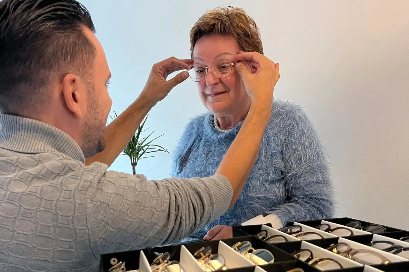 De opticien past bij een klant thuis een montuur, met de lade monturen ervoor